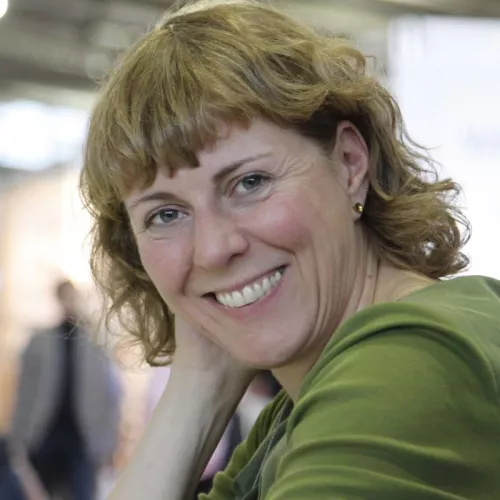 Andrea trägt eine grüne Bluse, lacht und schaut zur Seite in Richtung Kamera. Im Hintergrund eine Veranstaltung 
