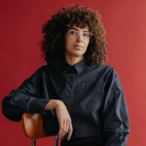 A brown skinned woman with curly dark hair and glasses, in a dark button-up shirt and dark pants, sits on a chair against a solid red background and looks at the camera.