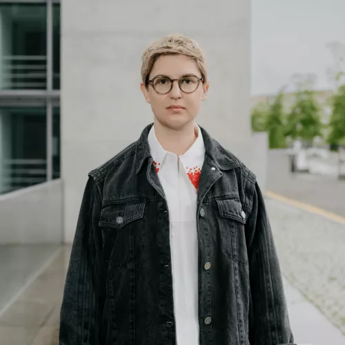 Porträtfoto von Charlotte Kunath. Charlotte steht vor einem modernen Gebäude im Freien, trägt eine dunkle Jeansjacke über einem weißen Hemd mit roter Stickerei am Kragen und eine runde Hornbrille. Kurzes blondes Haar.