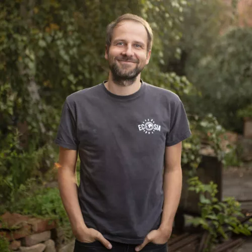 Christian Kroll steh in einem Wald, trägt ein kurzärmeliges dunkles T-Shirt und lächelt. Er hat kurze Haare und einen Bart. 