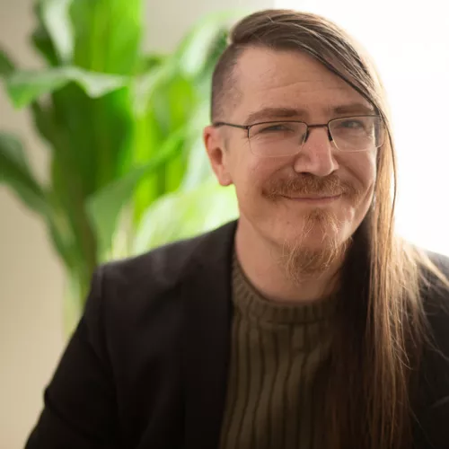 Christian Fein, mit Sidecut, Kinnbart, Brille, Rollkragenpullover und Sakko, blickt lächelnd in die Kamera. Im Hintergrund ist in der Unschärfe eine Zimmerpflanze zu sehen.