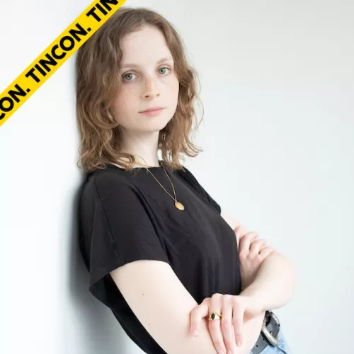 Corinna lehnt an einer weißen Wand und guckt in die Kamera. Ihr Oberkörper ist zu sehen. Sie hat offene dunkelblonde Locken, verschränkte Arme und trägt ein schwarzes T-Shirt.