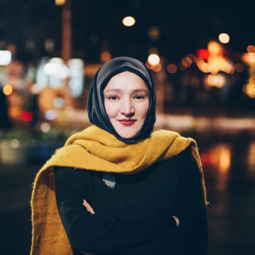 Eine Frau in Hijab und gelbem Schal verschränkt die Arme und lächelt in die Kamera.