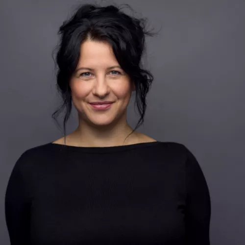 Henriette Lippold steht vor einem grauen Hintergrund und Blicks lächelnd in die Kamera. Sie trägt schwarze Haare und ein schwarzes Oberteil.