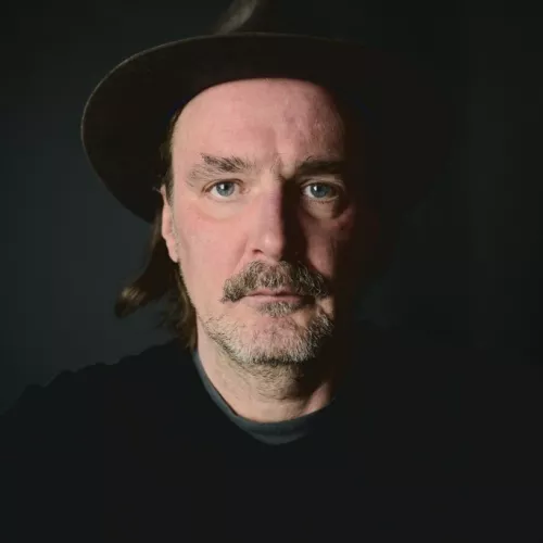 Ein (Selbst-) Portrait von Winson mit Hut vor dunklem Hintergrund. Das Licht kommt leicht seitlich rein, die rechte Gesichtshälfte ist dunkler als die linke.