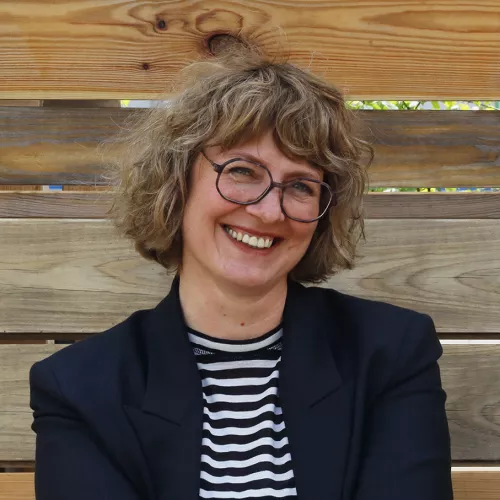 Prof. Daniela Hensel mit schwarz-weiß gestreiften Thirt und schwarzer Jacke sitzt vor einer Holzwand