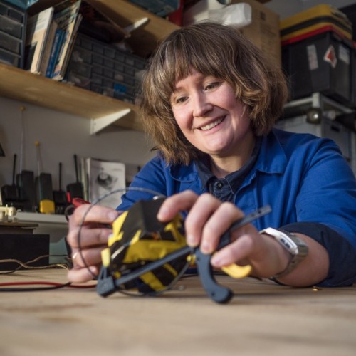 a smiling woman leaning on a workshop table using a multimeter to test a bunch of taped together 9 Volt batteries
