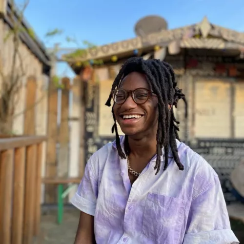 Dera is sitting at a rooftop table. He's wearing a loose purple-and-white button up and a big smile. His locs are tied up in a half do, and the blue sky and sun shine behind him