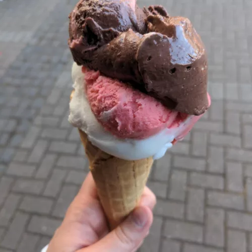Ein Bild von drei Eiskugeln in einer Eiswaffel, die von einer Hand gehalten werden. Die Sorten sind Zitrone, Erdbeere und Schokolade.