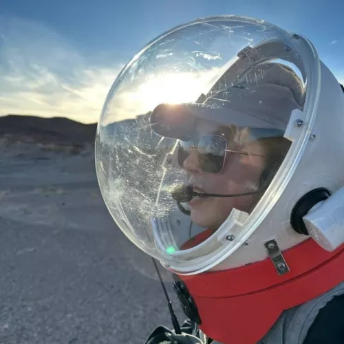 An analog astronaut wearing a simulation space suit with the sun shining through the helmet. She speaks through a radio mouthpiece in front of a desert mountain range.