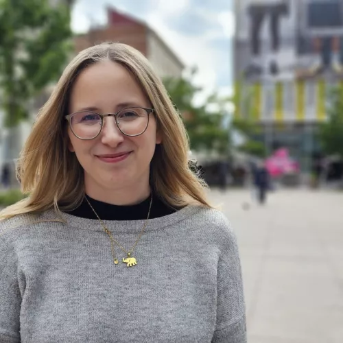 Porträt einer lächelnden Person im grauen Pullover mit goldener Kette. Die Person steht im Freien, im Hintergrund sind unscharf Gebäude und grüne Bäume zu sehen.