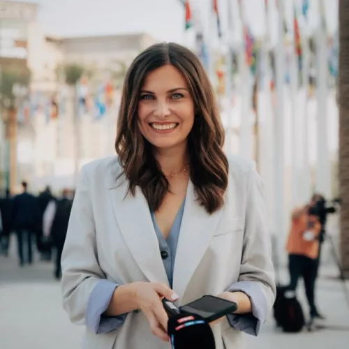 Clara Pfeffer auf dem Flaggen-Platz der COP28 in Dubai bei der Vorbereitung auf eine Live-Schalte mit einem Mikrophon und Handy in der Hand.