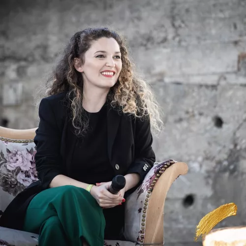 Aufnahme von Samira beim Female Future Festival 2022 in München. Sie ist eine junge Frau mit braunen Augen und langen brünett-blonden Kringellocken. Sie sitzt auf einem Sofa auf dem Podium, trägt ein schwarzes Top und Jackett und dunkelgrüne Stoffhose und hält ein Mikrofon im Schoß. Dabei schaut sie zum rechten Bildrand und lächelt jemanden außerhalb des Bildes an.