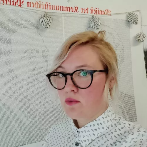 Veronika Kracher im Profil. Sie trägt eine Brille und eine weiße Bluse mit kleinen Turnschuhen darauf. Sie hat kinnlange, blonde Haare und steht vor einem Plakat mit Karl Marx und Friedrich Engels.