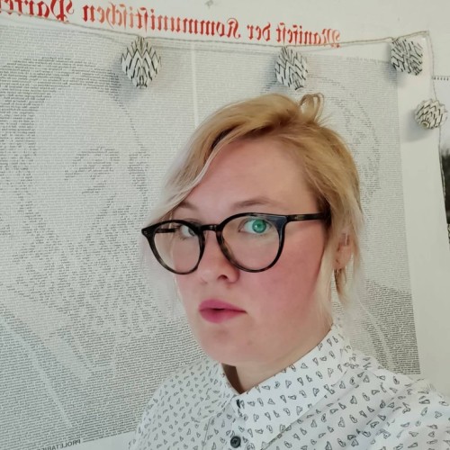 Veronika Kracher im Profil. Sie trägt eine Brille und eine weiße Bluse mit kleinen Turnschuhen darauf. Sie hat kinnlange, blonde Haare und steht vor einem Plakat mit Karl Marx und Friedrich Engels.