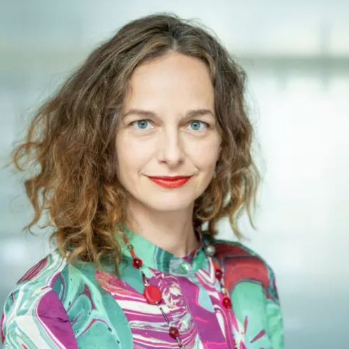Frau mit welligen braunen Haaren, buntem Oberteil und roten Lippen, lächelnd