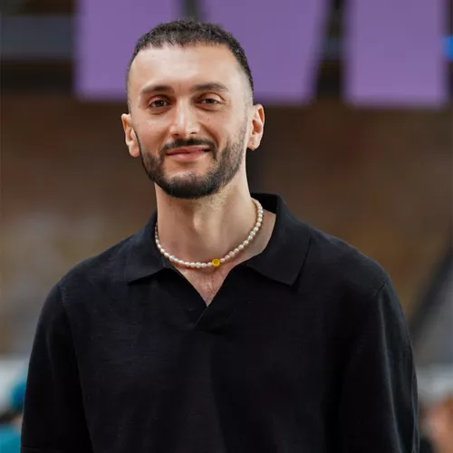 „Porträt von Marspet Movsisyan, einem Journalisten. Er hat kurze dunkle Haare, einen gepflegten Bart und trägt ein schwarzes Hemd mit Kragen. Um den Hals trägt er eine Perlenkette mit einem kleinen gelben Anhänger. Er lächelt leicht und steht vor einem unscharfen Hintergrund.“