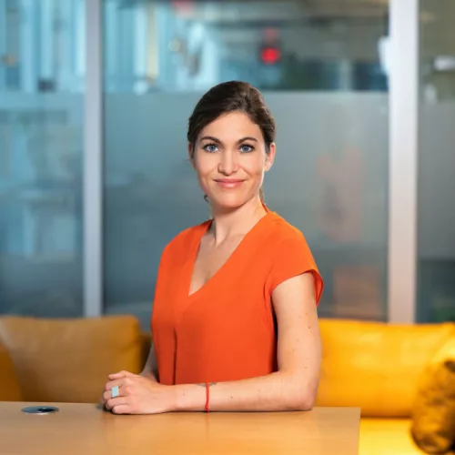 Foto von Verena Kauzleben in einem Büro. Dunkle Haare und seitlich angelehnt an einen Tisch. 