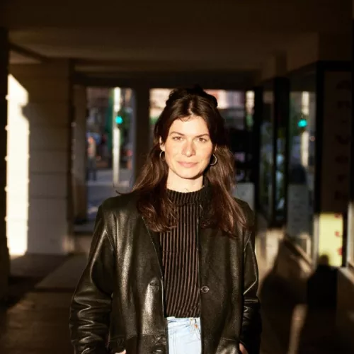Women with brown hair, black shirt and black jacket stands in a corridor and looks into the camera