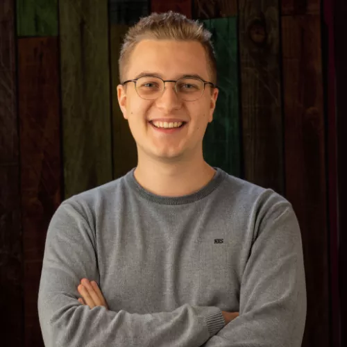 Foto von Tobias Bluhm in einem blauen Pullover und mit verschränkten Armen, er lächelt freundlich in die Kamera