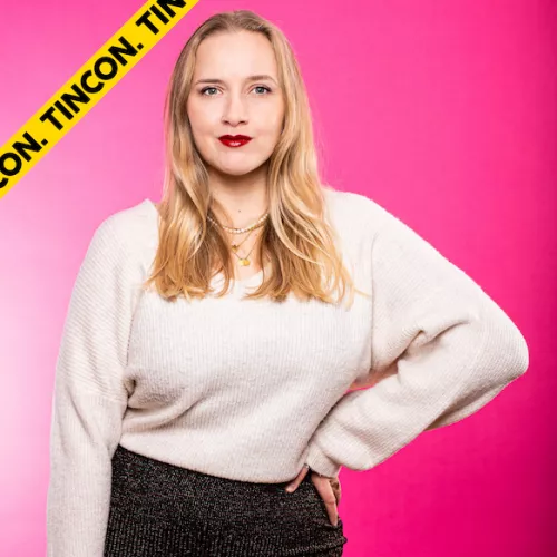 Frau Löwenherz trägt einen weißen Pulli. Sie steht und stützt ihren Arm an der Hüfte ab. Sie hat schulterlange, blonde Haare und guckt in die Kamera. Der Hintergrund ist pink.