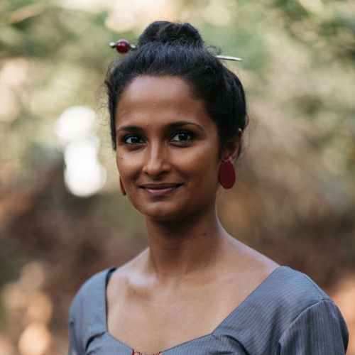 Gayatri, a dark skinned woman with black hair tied up in a bun, is pictured outdoors. She wears red round earrings, and smiles at the camera. She wears a grey striped top with red central pattern.
