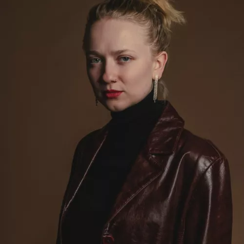 Mia Oberländer vor braunem Hintergrund. Sie trägt einen dunkelbraunen Ledermantel und roten Lippenstift.