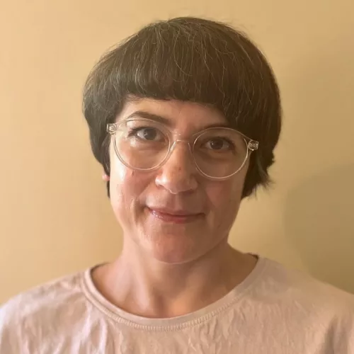 Frau mit dunkelbraunem kurzen Bob und heller Brille, hellem T-Shirt vor hellem Hintergrund