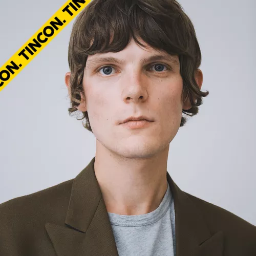 Hannes Vincent schaut in die Kamera. Das Foto ist ein Kopfausschnitt. er hat kurze braune Haare und trägt ein graues T Shirt mit braunem Blazer.