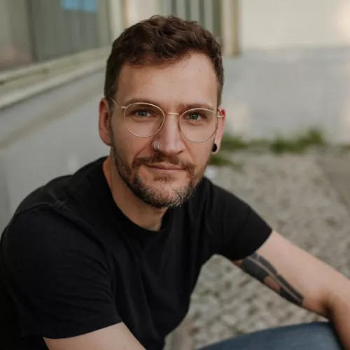 Mitte dreißigjähriger Mann mit kurzen hellbraunen Haaren und goldener Brille sitzt vor einem Türeingang.