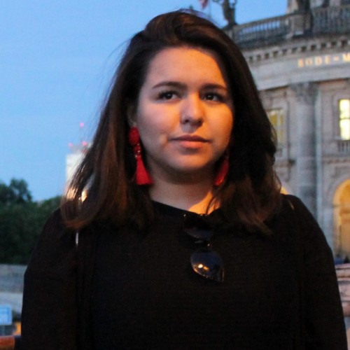 In the photo we see a young hispanic woman with brown eyes, medium-length hair and slightly tan skin, wearing red earrings and a black, long-sleeved pullover, standing in front of the Bode-Museum at dusk.