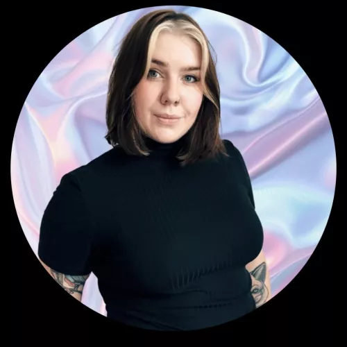 Das Bild zeigt eine Person mit mittellangem, glattem, braunem Haar, das vorne blonde Strähnen hat. Sie trägt ein schwarzes Oberteil mit einem hohen Kragen. Die Person schaut direkt in die Kamera und lächelt dezent. Der Hintergrund besteht aus einem glänzenden, pastellfarbenen Stoff, der in verschiedenen Lila-, Rosa- und Blautönen schimmert. Das Bild hat einen runden Rahmen mit schwarzem Rand.