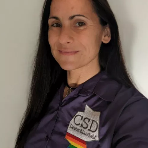 Weiblich gelesene Person mit langen dunklen Haaren trägt ein lila Hemd mit einer regenbogenfarbigen Deutschlandsilhouette mit dem Schriftzug "CSD Deutschland"