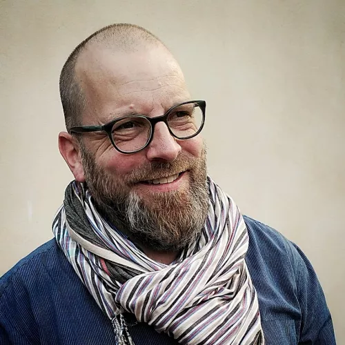 "Smiling bearded man with glasses and shaved head wearing a striped scarf, outdoor portrait."