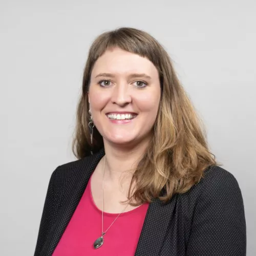 Das Foto zeigt das Portrait einer Frau, die vor einem weißen Hintergrund steht und in die Kamera lächelt. Sie trägt einen schwarzen Blazer und eine pinke Bluse.