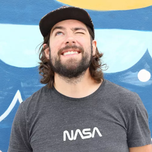 Portrait of Chris Spatschek in front of a Colourful Wall. He Wears a T-Shirt with NASA print and looks skywards, closing one eye. 