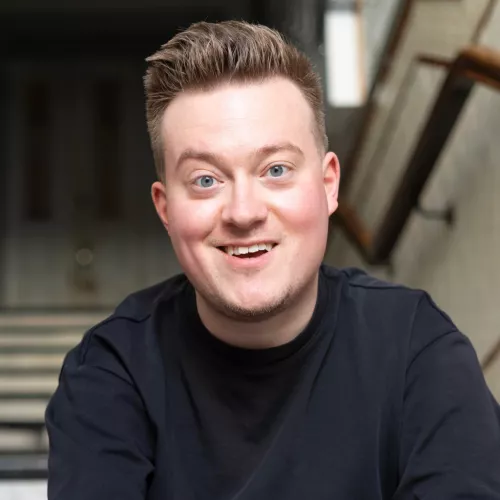 René Schaar trägt blondes kurzes Haar, ein schwarzes Shirt und lächelt in die Kamera. Er sitzt auf einer Treppe. 