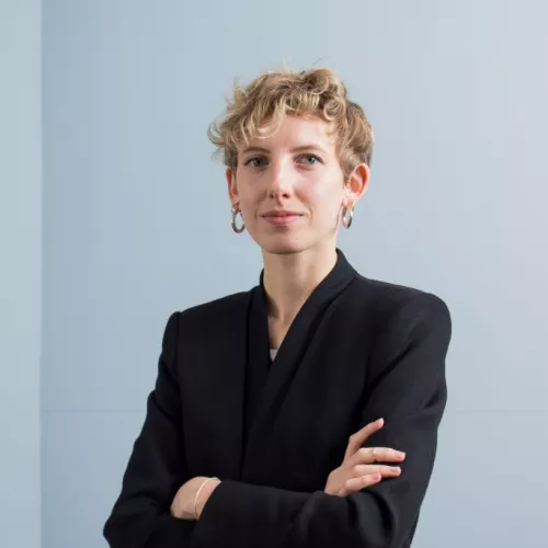 Bild von Julia Krickl, vor einer grauen Wand, stehend, Arme verschränkt vor der Brust.