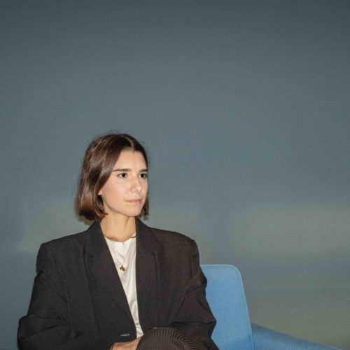 Weibliche Person im Seitenprofil trägt ein weißes Shirt und schwarzen Blazer und sitzt auf einem blauen Sessel. 