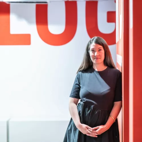 Maureen Ekizoglu lehnt mit ihrer linken Schulter gegen eine rote Wand. Sie lächelt freundlich in die Kamera.