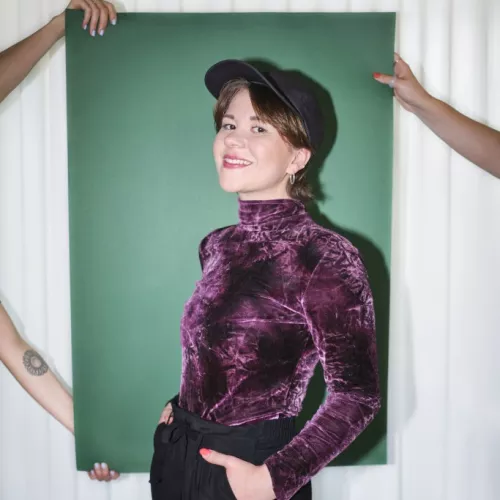 Das Bild zeigt Anna-Karina Vollmer, eine Frau mit einer schwarzen Cap, die seitlich steht und ihre Hände in die Taschen der schwarzen Stoffhose gesteckt hat. Sie trägt ein lilafarbenes Samtoberteil mit langen Ärmeln und Rollkragen. Sie lächelt in die Kamera und steht vor einem grünen Plakat, welches von drei Händen hinter ihr gehalten wird. Hinter dem Plakat befindet sich ein weißer Vorhang.