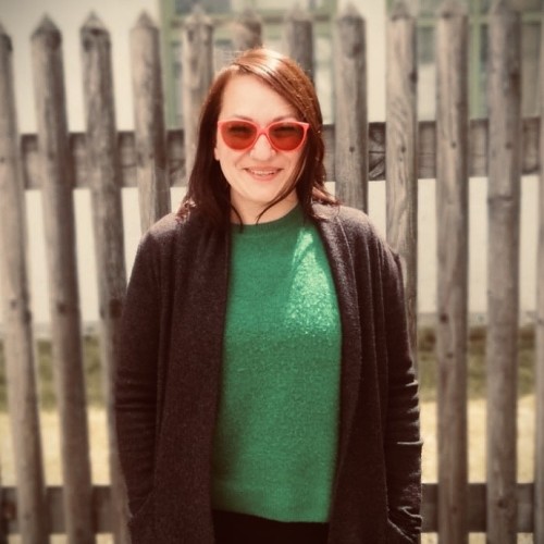 Karolina Capasso im grauen Mantel mit grünem Pullover und roter Brille vor einem Holzzaun.