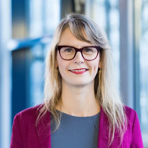 Portraitfoto einer Frau mit blonden, etwas über Schulterlangen Haaren, roter Brille mit grauem Oberteil und auberginefarbenem Blazer vor verschwommenem Fensterhintergrund