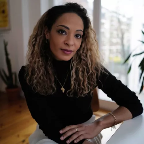 Kemi Fatoba sitzt neben einem Tisch, an den sie sich anlehnt, und blickt in die Kamera. Sie trägt ein schwarzes Oberteil und einen weißen Rock.