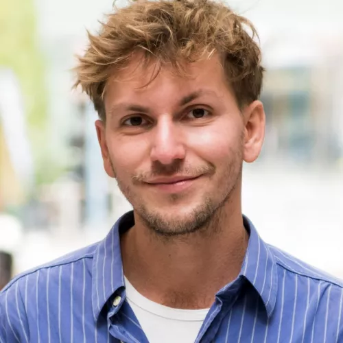 Wir sehen eine weiße Person mit welligem dunkelblondem Haar. Die Person trägt ein blau gestreiften Hemd mit einem weißen T-Shirt darunter.