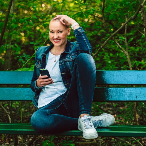 draußen: Laura in Jeans und Jeansjacke lehnt lässig auf einer Bank, mit blondem Buzzcut und Handy in der Hand lächelt sie in die Kamera