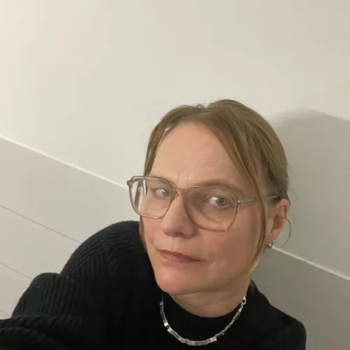 Zu sehen ist eine Frau mittleren Alters. Sie hat eine viereckige modische Brille auf und die längeren hellen Haare in einem Pferdeschwanz zusammengebunden. Sie trägt ein schwarzes Oberteil und eine silberne Kette. Ihr Blick in die Kamera ist neugierig. Aufgenommen ist das Foto in einem weißen Raum. Die Haltung lässt darauf schließen, dass es ein Selfie ist.