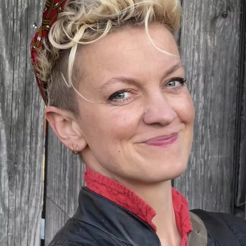 Lillian Rosa, kurze blonde Haare und rote Käppi, steht vor einer Holzwand und lächelt in die Kamera