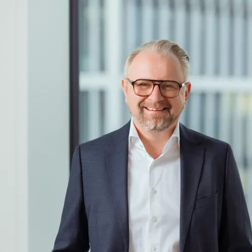 Nikolaus hat blonde Haare, trägt ein dunkles Jackett und Brille. 