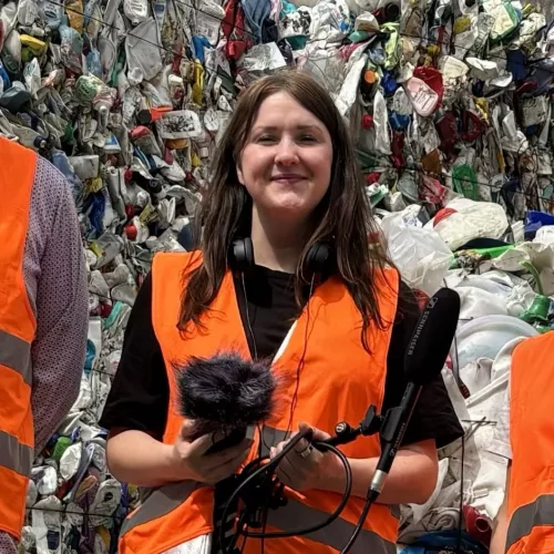Maike Klein steht in einer Warnweste mit Aufnahmegerät und Mikrofon in der Hand vor meterhoch sortenrein aufgetürmten Kunststoffverpackungen